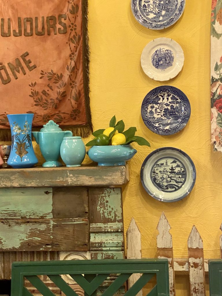 Fireplace mantle with patina, picket fence indoors, blue and white china on yellow wall